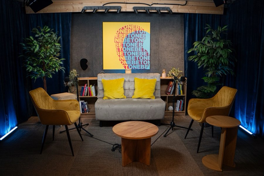 Studio 1 from the front in blue light: a grey sofa, two mustard armchairs, two microphones and a round wooden table.
