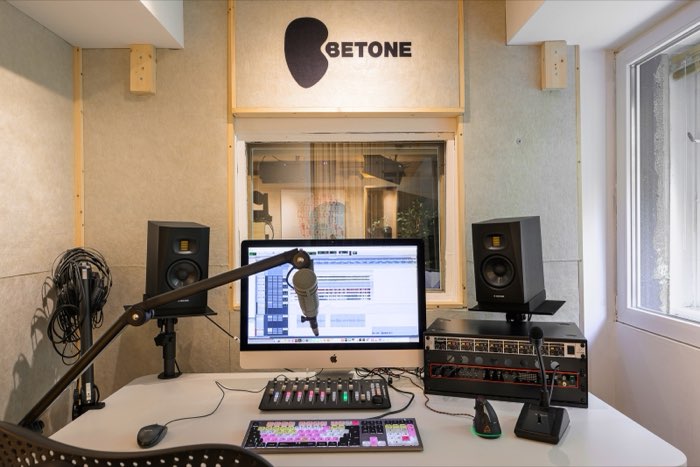 Studio 2 from the front: a computer, two monitor speakers and a microphone; a Betone logo above, with Studio 1 visible through the window.